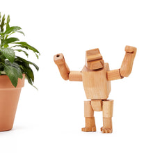 Hanno Jr., a small wooden gorilla toy, stands with arms raised near a potted plant. The gorilla is made of beautiful wood and assembled by elastic string.