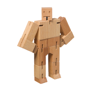 A wooden robot stands with hands on hips and looks at the camera. The robot is made of chunky rectangular pieces and has a simple face etched into the head.