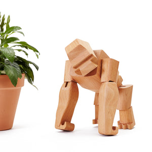 Hanno, a wooden gorilla toy, stands near a potted plant. Hanno is made from sculpted pieces of wood attached by strong elastic string.