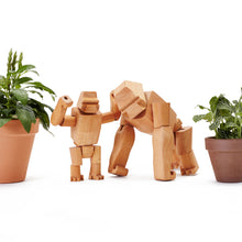 Two wooden gorillas stand between potted plants. The larger gorilla stands on all-fours, the smaller gorilla stands with both hands held above it's head.