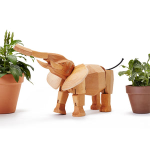 Wooden elephant toy stands between two potted plants. The elephant is standing with trunk raised.