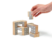 A hand places a grey wooden block onto other blocks to create a building shaped form. The wooden blocks are grey with white painted windows.