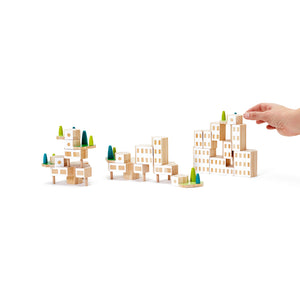 A hand places a white painted wooden block on a stack of blocks that looks like a building. Two other small block buildings are shown in the foreground.