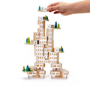 A hand places a wooden block at the top of a tall block tower. The tower looks like a white skyscraper with cantilevered platforms containing trees.