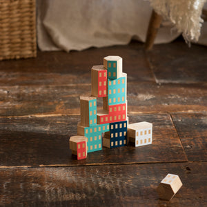 Brightly colored blocks are stacked in the shape of skyscraper and sit on a rustic wooden floor. The blocks are painted red, white, light blue, and navy blue, and appear to have windows, making the blocks look like buildings in a city.