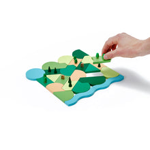 A hand places a wide, flat wooden block into a series of other wooden blocks that look like a park. The blocks are painted a variety of shades of light brown, green, and blue.