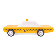 A side view of the a wooden taxi cab. The bottom half of the car is yellow with traditional black checkerboard decoration, the top is white and sports a taxi sign on the roof.