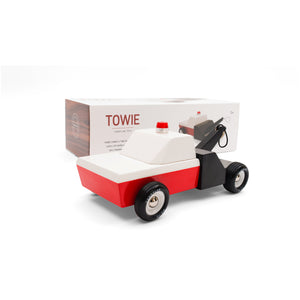 A wooden toy tow truck in front of its packaging. The truck is red and white, with a black rear end. An orange safety light is centered on the roof.