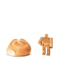 A micro sized wooden robot stands next to a dinner roll. 