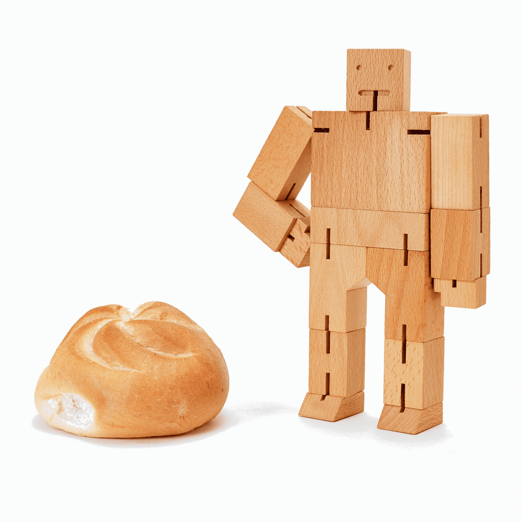 A medium sized wooden robot toy stand next to a dinner roll. The robot is made of wooden rectangular blocks and assembled with elastic string.