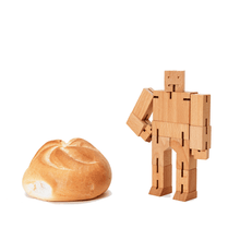 A small wooden robot stands near a dinner roll. The robot has it's hand on the right hip.