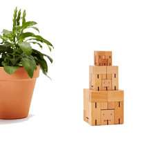 A tower of wooden robots formed into cubes is pictured next to a potted plant. Each robot is a different size.