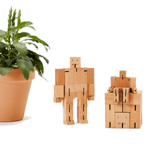 A group of three wooden robot toys are pictured next to a potted plant. The robots represent the three sizes of cubebot, the medium is a cube and is used as a seat for the micro, while small stands nearby.