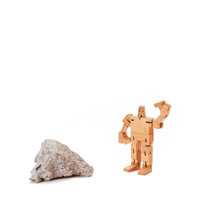 A micro sized wooden robot named Guthrie stands next to a rock. 