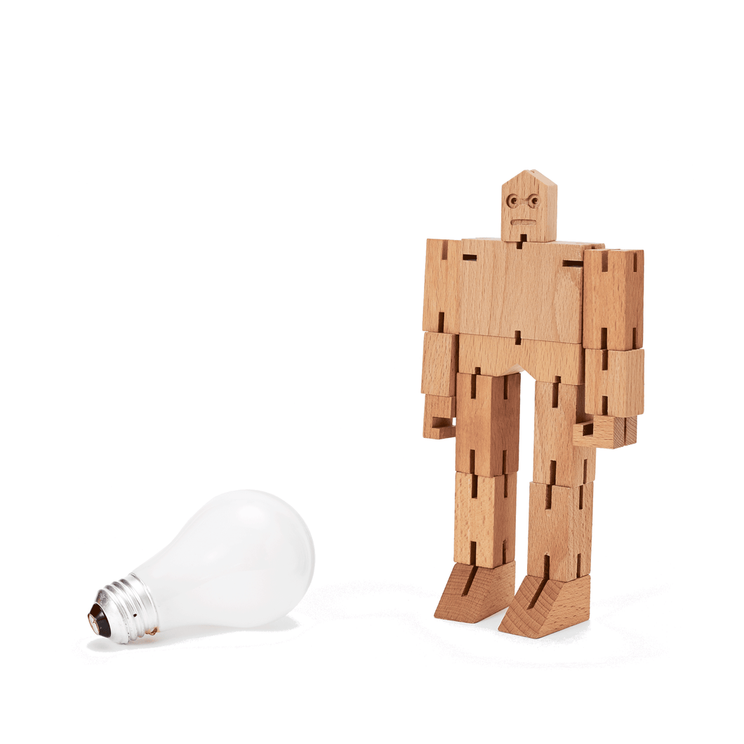 A small wooden robot is pictured next to an incandescent light bulb. The robot is almost twice as tall as the lightbulb.