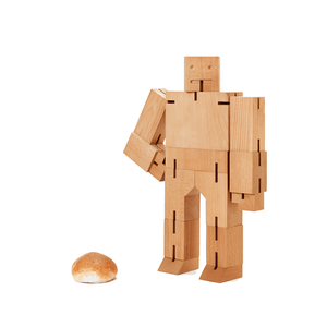A giant wooden robot stands next to a dinner roll for scale. The robot has a hand on it's right hip, and is made of large wooden blocks joined by elastic.